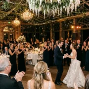 Bride and groom dancing in a candlelit barn venue under fairy lights while guests clap and a live band plays